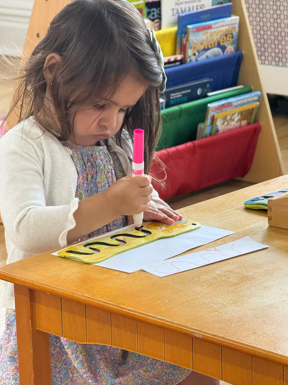A child tracing curves with a marker, Spanish picture books on the shelf behind her